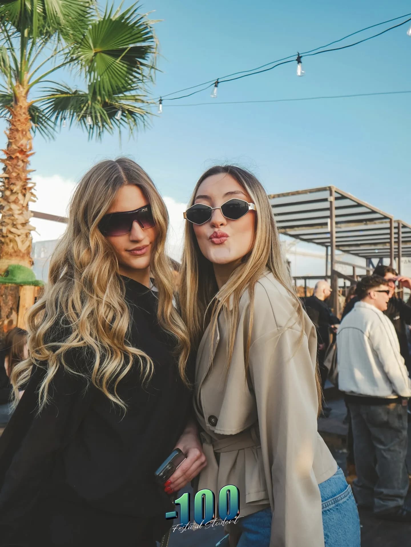 Due ragazze sorridono sotto una palma, con le lucine accese al tramonto.